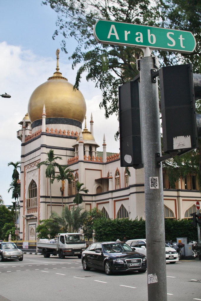 Uma cena de rua mostrando a cúpula dourada da Mesquita do Sultão com carros na rua, sob uma placa verde "Arab St" em Singapura.