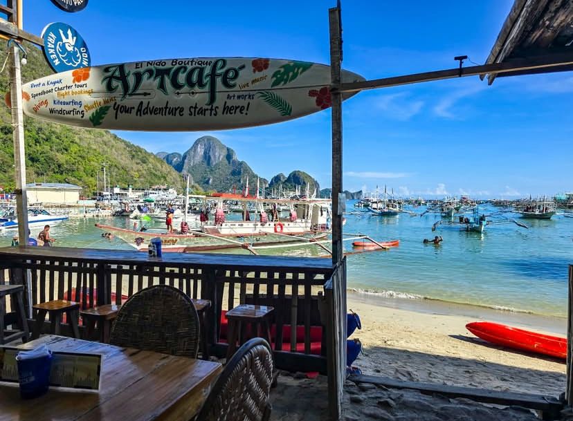 Vista de um café na praia mostrando barcos na água, montanhas ao longe e um céu azul claro.