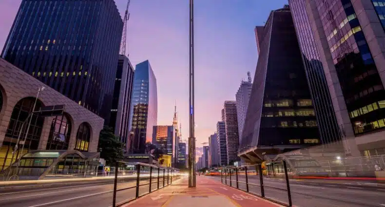 Vista ampla da Avenida Paulista com arranha-céus modernos em ambos os lados, uma ciclovia no centro e um pôr do sol ao fundo. Imagem para ilustrar post sobre pontos turísticos de São Paulo.