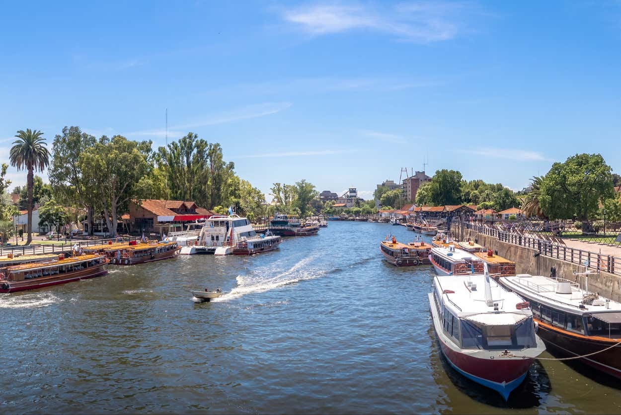 Uma cena de rio no Delta do Tigre, um dos pontos turísticos de Buenos Aires, movimentado com vários barcos atracados ao longo de uma costa arborizada sob um céu azul claro.