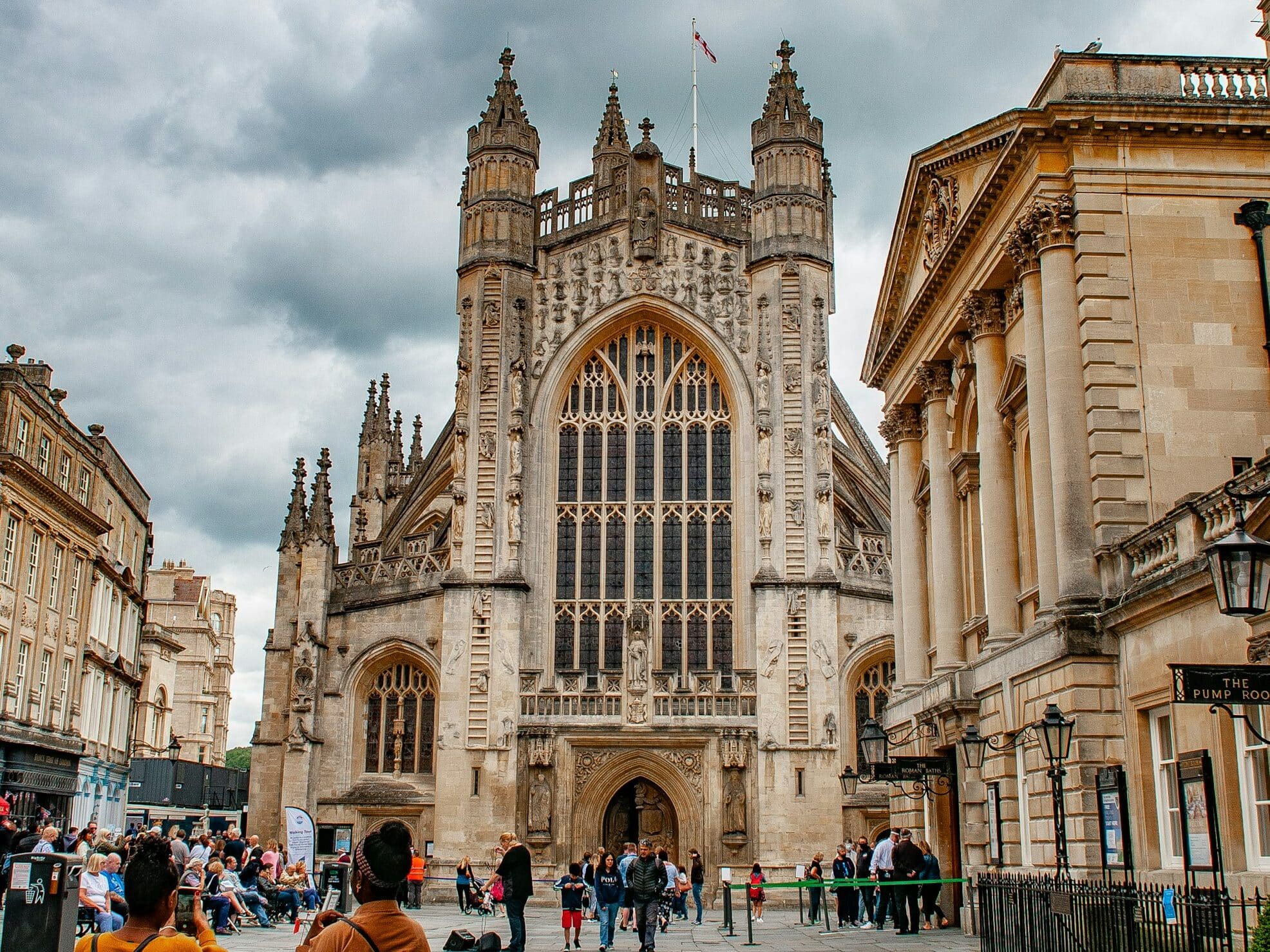Pessoas tirando fotos da Abadia de Bath, uma grande catedral histórica com torres altas e uma fachada ampla sob um céu nublado. Foto para a lista de o que fazer em Bath.