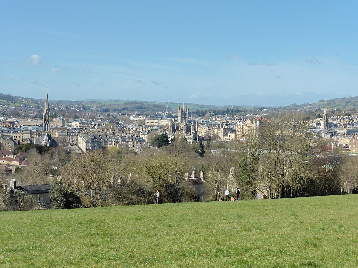 Uma colina gramada com vista para a paisagem urbana de Bath, com edifícios históricos, torres e árvores sob um céu azul claro. A vista faz parte da Bath Skyline Walk, perto de Bathwick Hill.