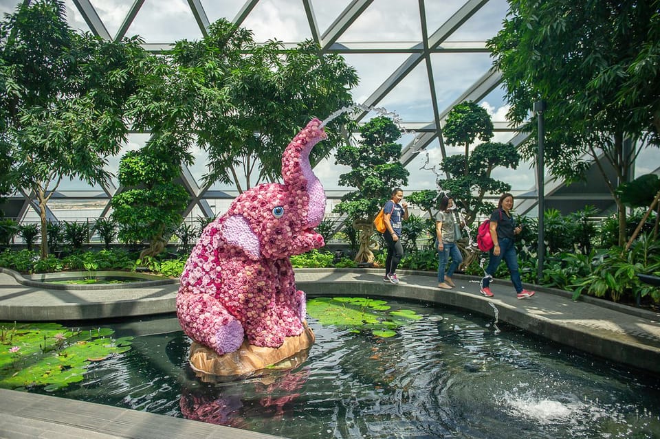 Uma escultura de elefante com flores rosa fica em um lago dentro de uma cúpula geodésica, cercada por vegetação e três pessoas caminhando.