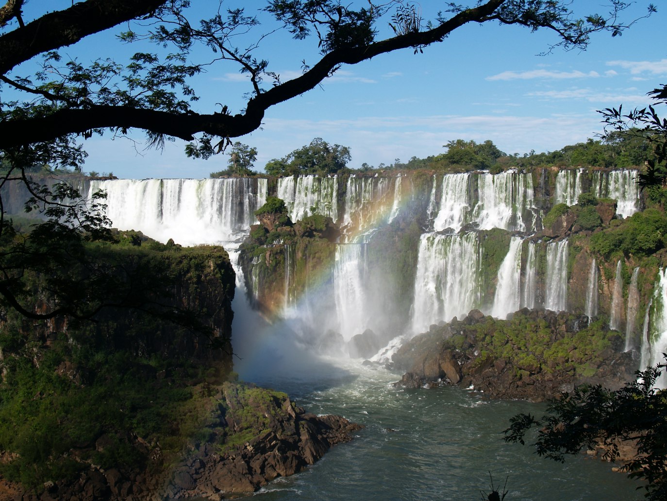 Cataratas do Iguazú despencam sobre penhascos verdejantes e exuberantes em um rio. Um arco-íris se forma em meio à névoa sob um céu azul, emoldurado por galhos de árvores.