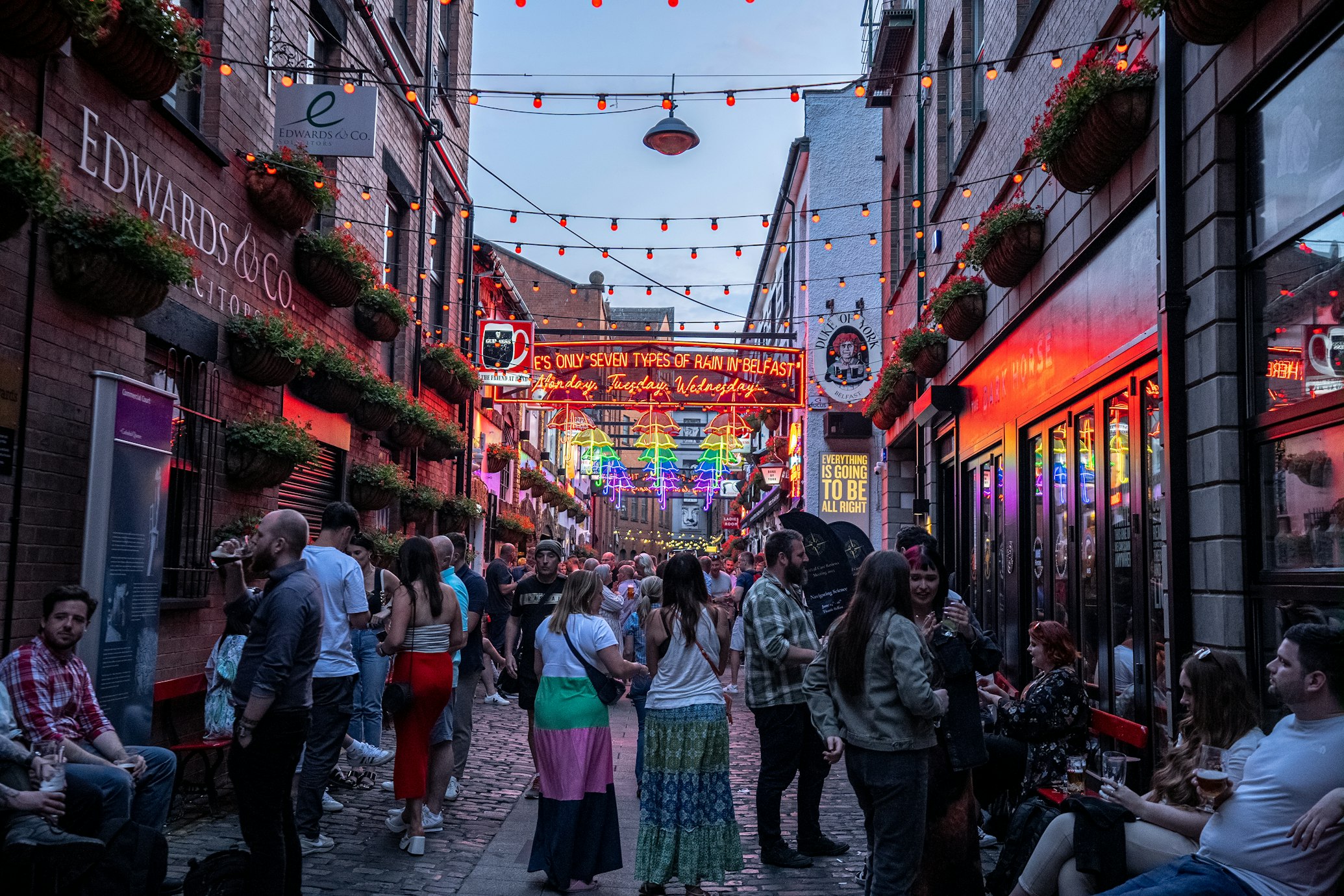 Um beco vibrante na Cathedral Quarter com luzes coloridas, lotado de pessoas aproveitando uma noite de reunião e socializando perto de placas iluminadas. Foto para a lista de o que fazer em Belfast.