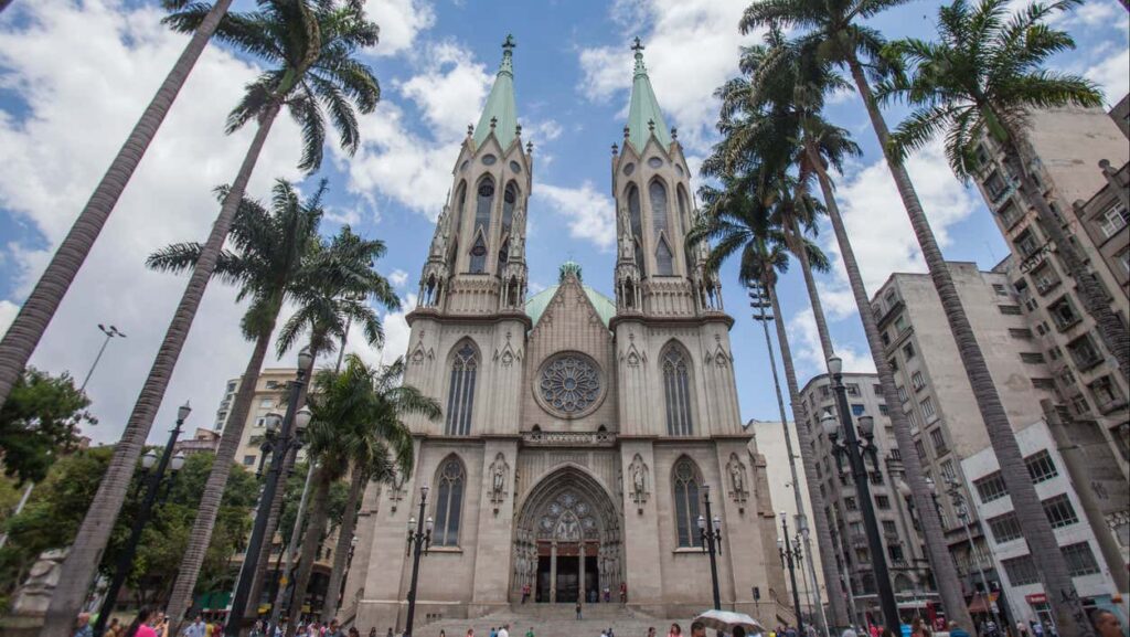 Catedral de Sé com torres gêmeas e uma rosácea, cercada por altas palmeiras em um dia ensolarado com céu parcialmente nublado.