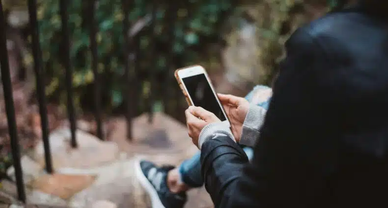Pessoa sentada em degraus de pedra ao ar livre, segurando um smartphone, com vegetação desfocada e grades de metal ao fundo. A imagem ilustra o post sobre eSIM ou SIM físico.