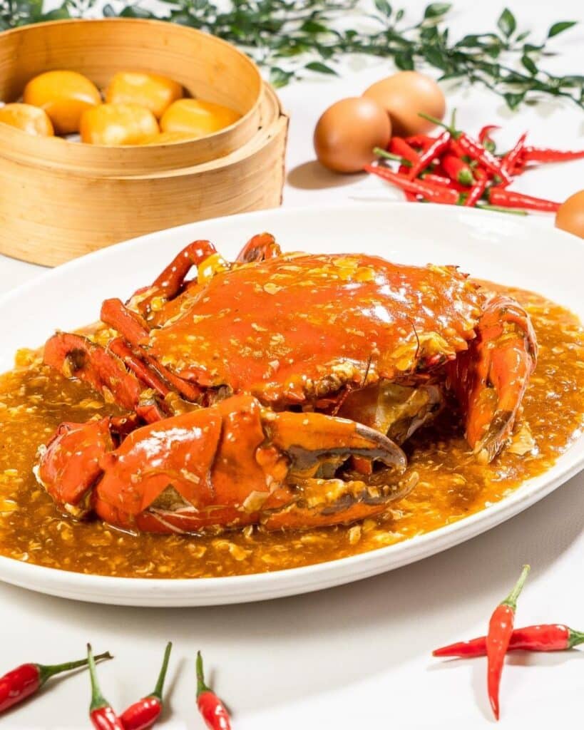 Um prato de caranguejo com molho apimentado, cercado por pimentas vermelhas, ovos e uma panela de vapor de bambu com pães ao fundo. É no restaurante Palm Beach Seafood em Singapura.