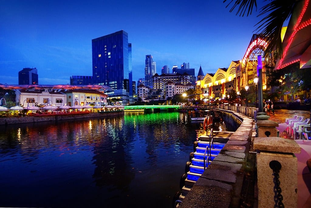 Vista noturna do Riverside Point em Clarke Quay com edifícios iluminados refletindo na água; o horizonte da cidade visível ao fundo. Representa o que fazer em Singapura.