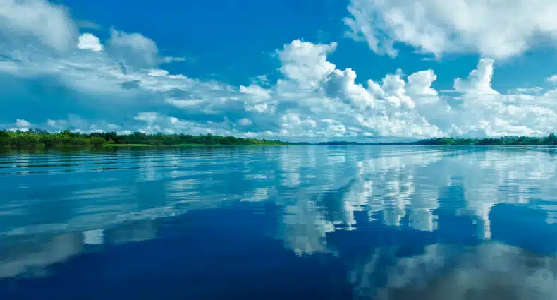 Rio Demerara calmo com céu azul e nuvens refletidas na água, ladeado por árvores verdes ao longe. Foto tirada na Guiana.