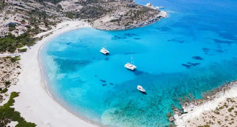 Vista aérea de uma baía serena com águas azul-turquesa, praia de areia, colinas rochosas e três veleiros ancorados perto da costa. A imagem ilustra o post sobre o que fazer na Ilha de Milos.