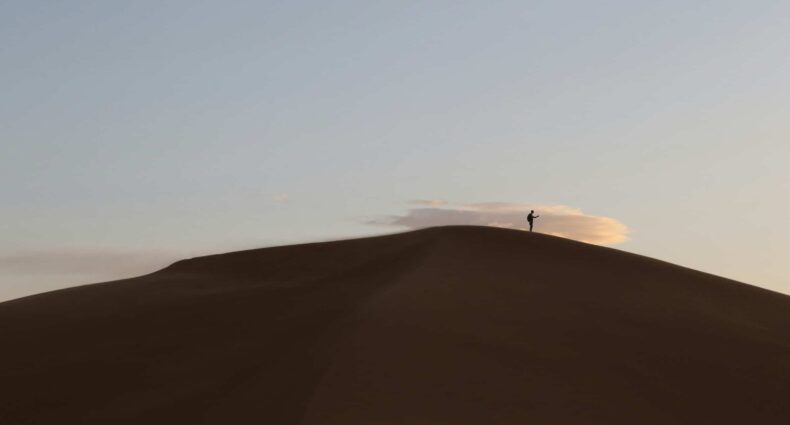 Uma pessoa está sozinha no topo de uma duna de areia no deserto do Saara sob um céu claro com uma pequena nuvem ao fundo, que pode ser visitado com o eSIM Marrocos.
