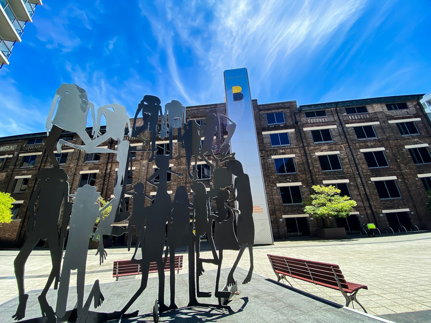 Esculturas de metal de figuras humanas ficam em um pátio com um prédio de tijolos e uma alta estrutura de vidro ao fundo.