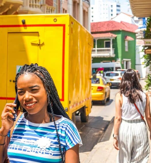 Uma mulher com tranças sorri enquanto fala ao telefone em uma rua ensolarada da cidade, cheia de carros e um caminhão amarelo. A imagem ilustra o post sobre eSIM Colômbia.