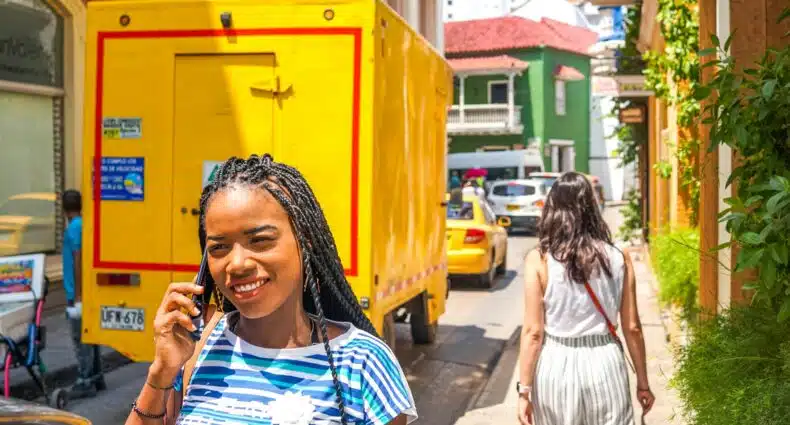Uma mulher com tranças sorri enquanto fala ao telefone em uma rua ensolarada da cidade, cheia de carros e um caminhão amarelo. A imagem ilustra o post sobre eSIM Colômbia.