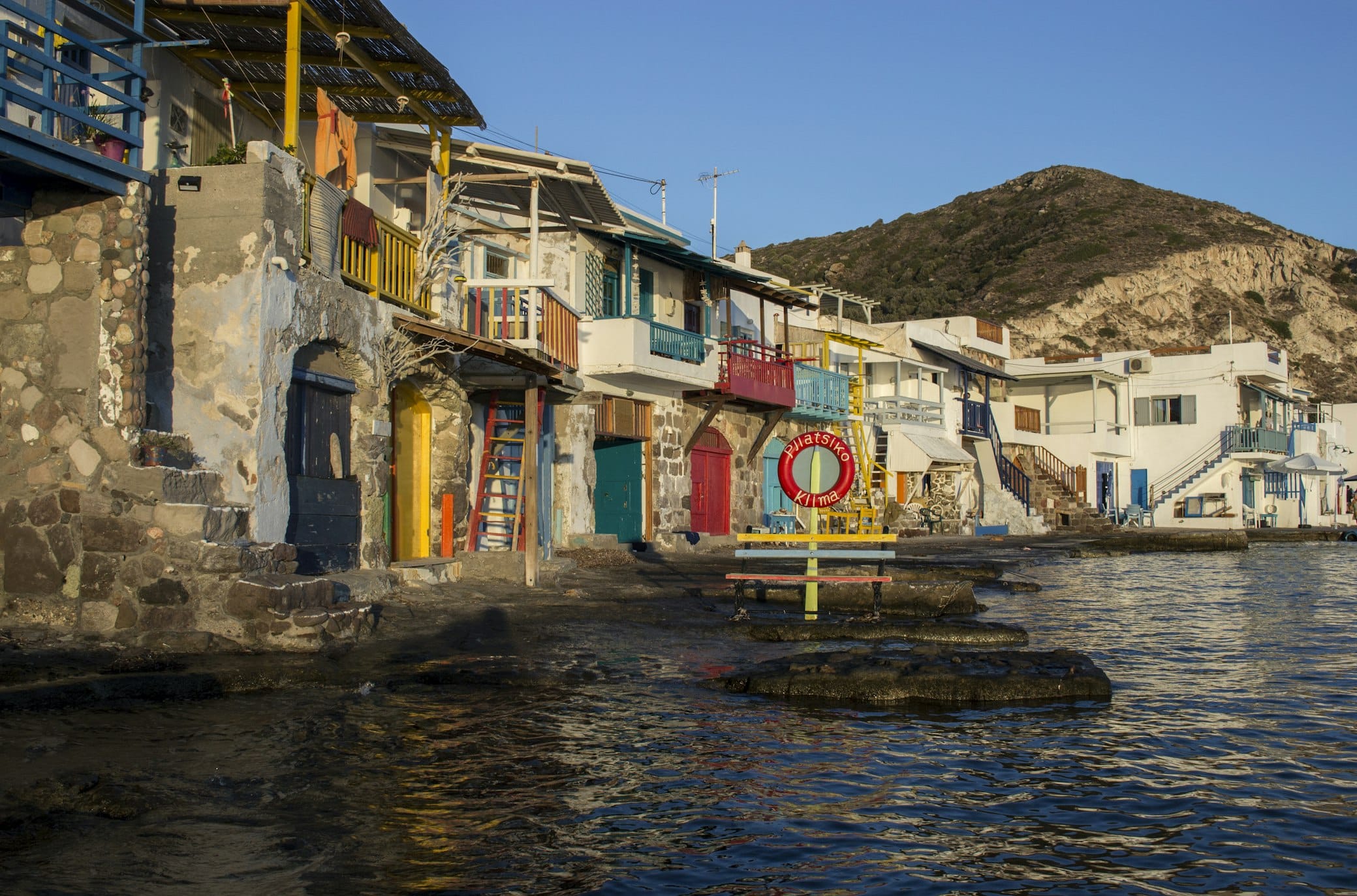 Casas coloridas com varandas se alinham à beira-mar, com uma montanha ao fundo sob um céu azul claro.
