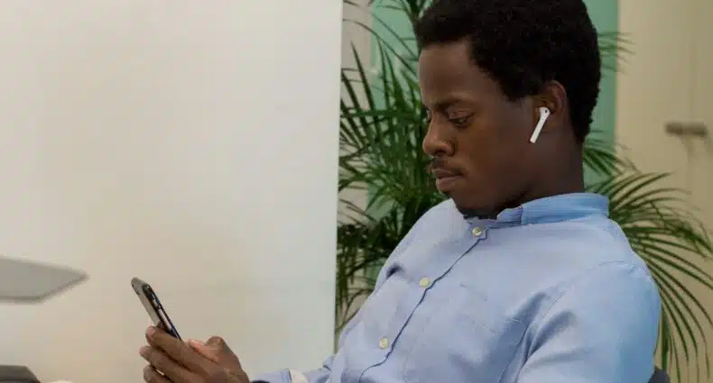 Um homem usando fones de ouvido está sentado em uma mesa e olha para o telefone, com uma planta e uma porta ao fundo em Luanda. Para ilustrar o post de eSIM Angola.