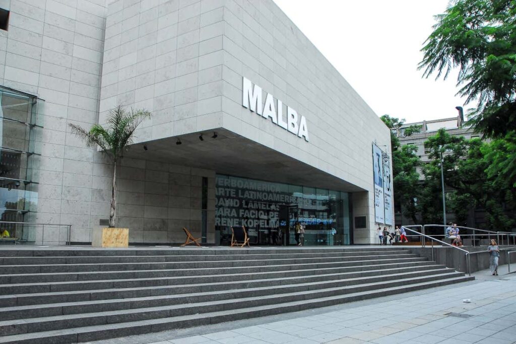 Entrada do MALBA com grande fachada de pedra, escadas que levam para cima, placa "MALBA", portas de vidro e árvores próximas. Pessoas à distância.