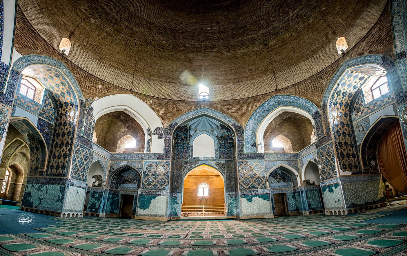 Interior da Masjed-e Kabud com azulejos azuis ornamentados, janelas em arco e um grande teto abobadado sobre um piso acarpetado estampado. Imagem para ilustrar post sobre o que fazer no Irã.