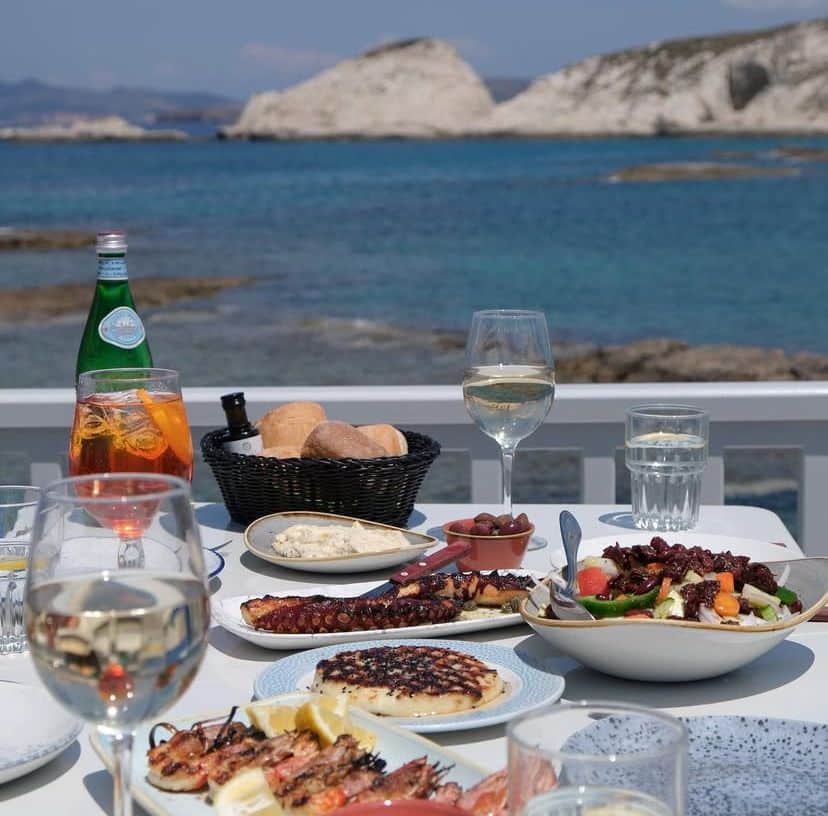 Uma mesa com frutos do mar, saladas e bebidas tem vista para o mar, com formações rochosas e águas azuis cristalinas.