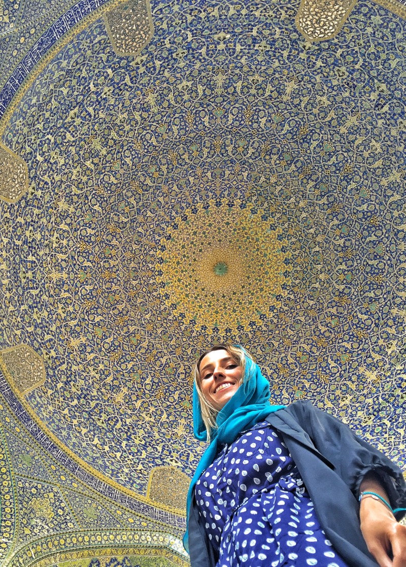 Uma de nossas colunistas com um lenço azul na cabeça, em pé sob uma cúpula de mosaico colorido e ornamentado na Mesquita Shah, sorrindo e olhando para cima. Imagem para ilustrar post sobre o que fazer no Irã.
