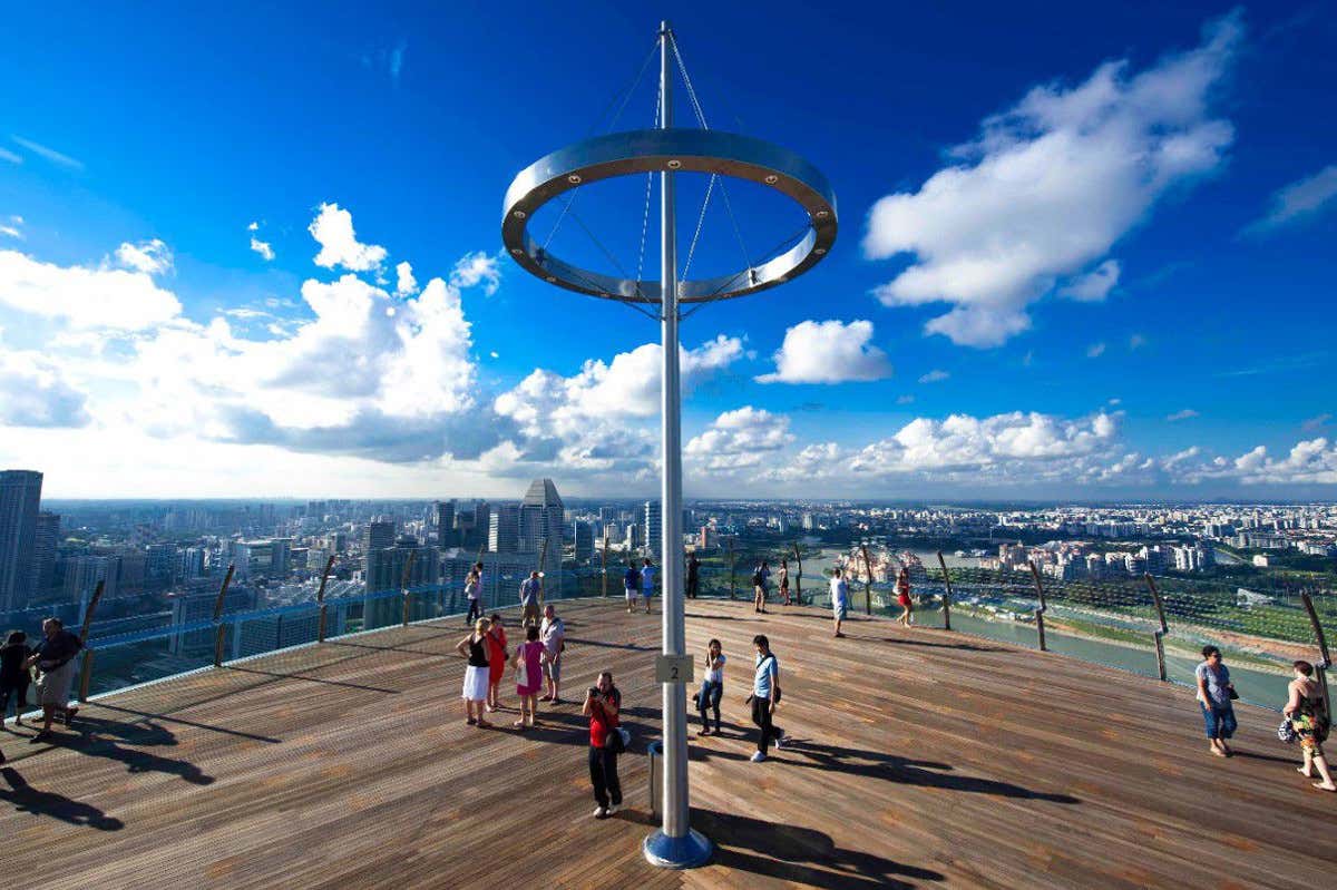 Pessoas caminhando em um grande mirante do Hotel Marina Bay Sands com uma estrutura de poste central; horizonte da cidade e céu azul com nuvens ao fundo. Ilustra o que fazer em Singapura.