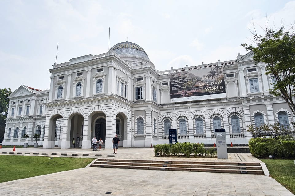Um grande edifício branco em estilo colonial, com janelas em arco e uma cúpula, com uma grande faixa de um lado, cercado por vegetação. É o Museu Nacional de Singapura.