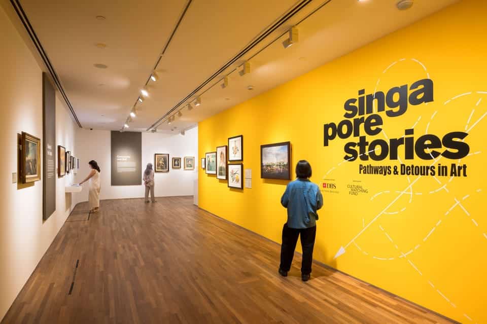 Uma galeria com pessoas vendo arte. Uma parede amarela exibe o texto "Singapore Stories: Pathways & Detours in Art". Representa o que fazer em Singapura.