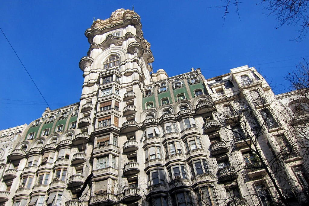 Edifício ornamentado de vários andares com varandas arredondadas, fachada verde e branca e uma torre com uma grande cúpula contra um céu azul.