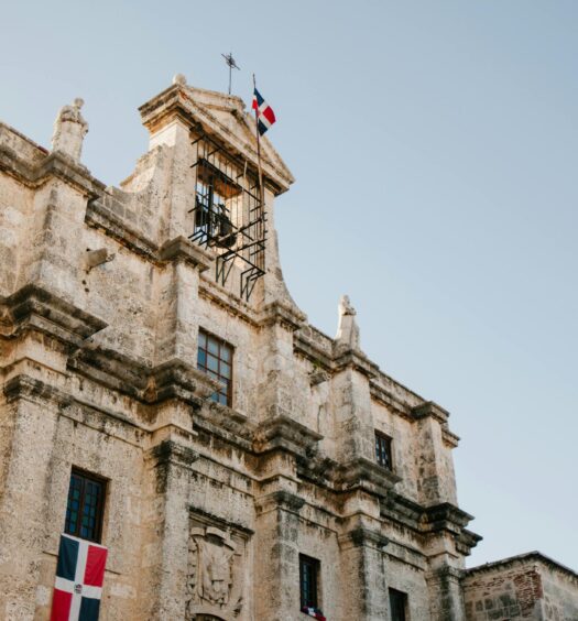 Edifício histórico de pedra com estátuas decorativas e várias janelas, com uma bandeira no topo e outras penduradas abaixo.