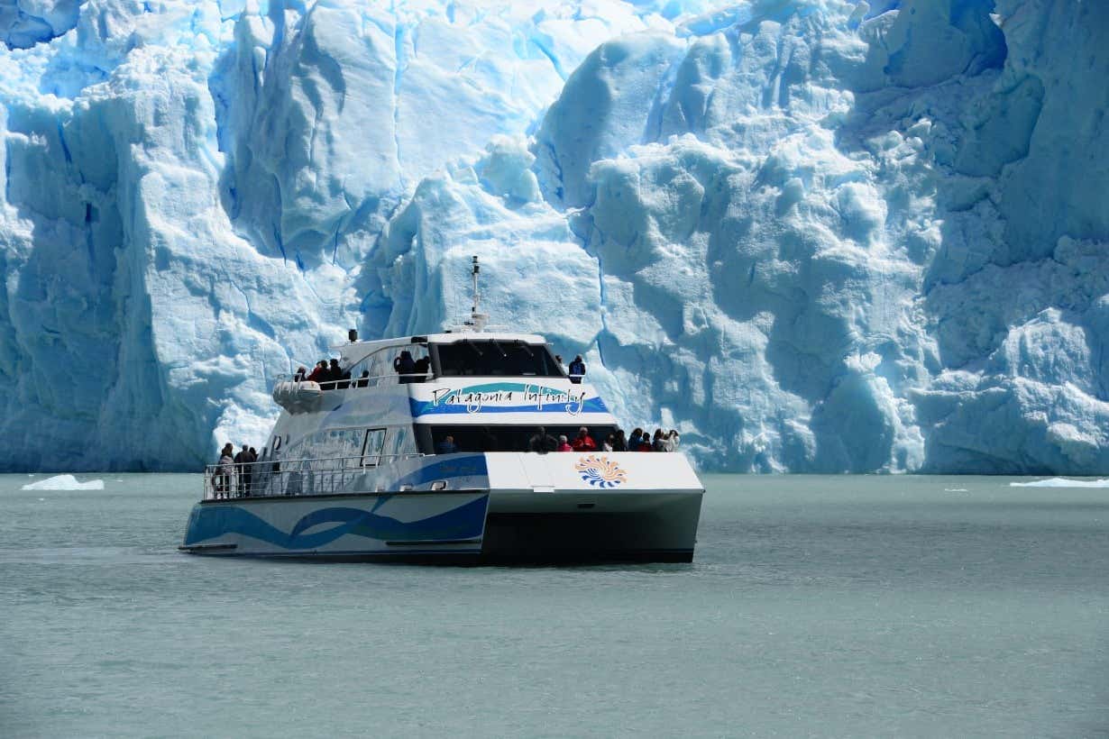 Um barco turístico no Parque Los Glaciares com passageiros se aproxima de uma enorme geleira, flutuando em águas calmas e azul-turquesa sob a luz do dia.