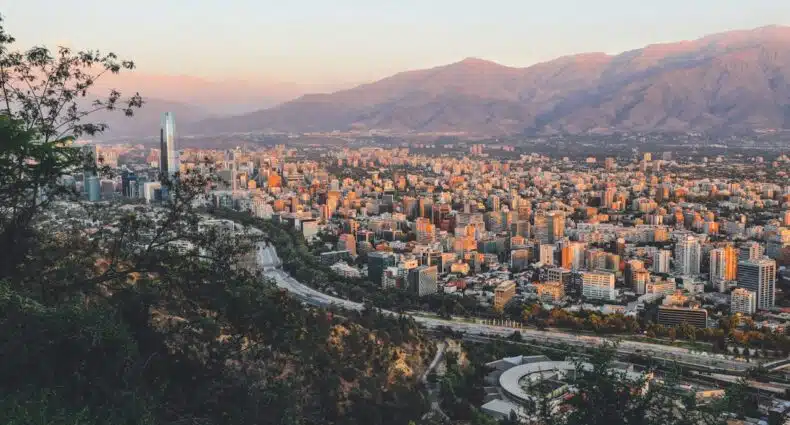 Vista panorâmica de uma paisagem urbana com vários edifícios, cercada por montanhas sob um céu claro durante o pôr do sol. A imagem ilustra o post sobre eSIM Santiago.