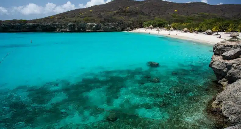 Uma baía azul-turquesa com uma costa rochosa, praia de areia e uma colina ao fundo, sob um céu parcialmente nublado. A imagem ilustra o post sobre eSIM Curaçao.