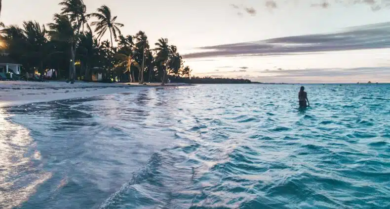 Uma pessoa está em águas rasas do oceano ao pôr do sol, com palmeiras alinhadas na praia e ondas quebrando suavemente na costa. A imagem ilustra o post sobre eSIM Rapública Dominicana.