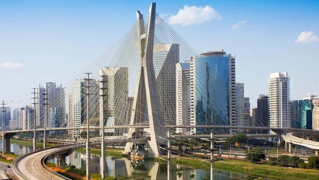 Ponte estaiada com duas torres inclinadas sobre um rio, cercada por arranha-céus modernos e estradas sob um céu azul. Imagem para ilustrar post sobre pontos turísticos de São Paulo.