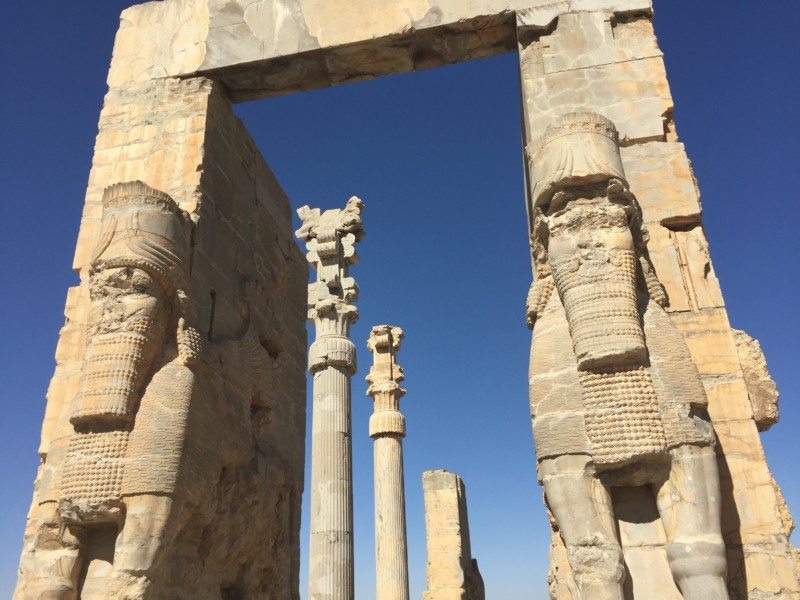 Portão de Todas as Nações com colunas altas e esculturas estilizadas de figuras humanas em contraste com um céu azul claro.