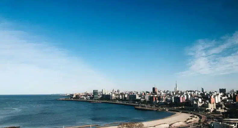 O horizonte de uma praia urbana em Montevidéu, com edifícios ao longo da costa e um céu azul com nuvens dispersas. A foto está no post de eSIM Montevidéu.