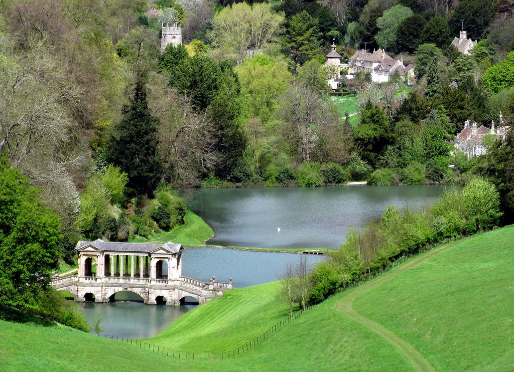 Uma vista panorâmica de jardins paisagísticos com uma ponte palladiana sobre um lago, cercados por árvores e casas distantes sob um céu claro. Essa é uma opção imperdível de o que fazer em Bath.