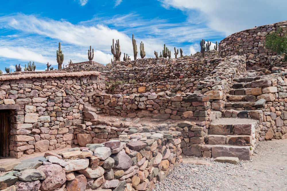 Antigas ruínas de pedra com degraus em uma paisagem desértica, cercadas por cactos sob um céu azul com nuvens dispersas.