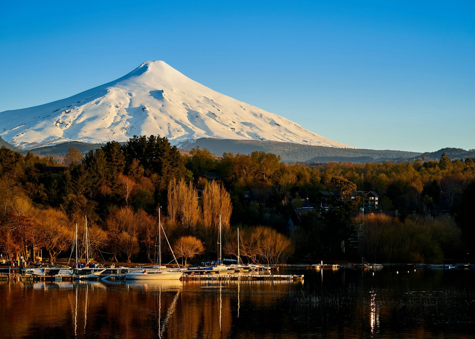 Vulcão coberto de neve com veleiros atracados em um lago calmo em primeiro plano, sob um céu azul claro. A imagem ilustra o post sobre eSIM Chile.