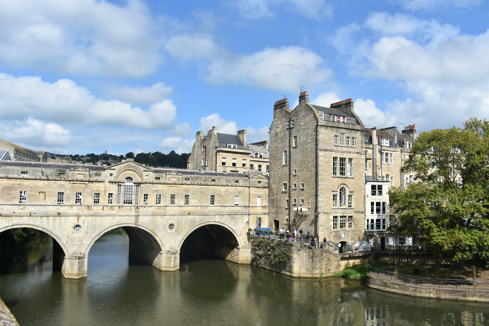 Pulteney Bridge, ponte de pedra e prédios históricos sobre o rio Avon que está calmo e sob um céu azul parcialmente nublado. Árvores e colinas luxuriantes ao fundo. Foto para a lista de o que fazer em Bath.