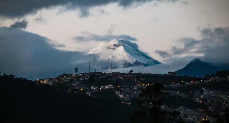 Uma montanha na cordilheira dos Andes coberta de neve ergue-se acima de uma cidade iluminada, vista sob um céu nublado ao anoitecer ou ao amanhecer. Foi tirada em Quito, no Equador.