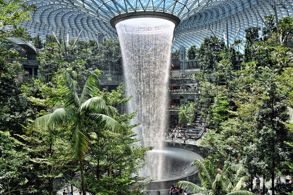 Grande cachoeira interna cercada por plantas tropicais sob uma cúpula de vidro, com pessoas observando de vários níveis. É a Rain Vortex em Singapura.