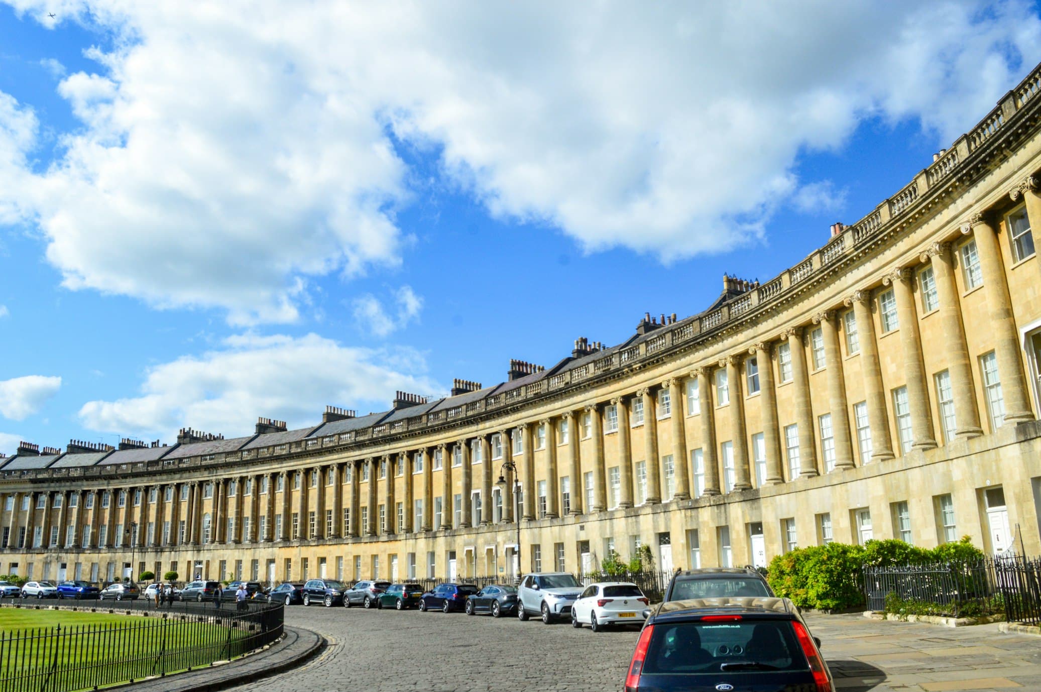 Royal Crescent, uma fileira de casas históricas com fachadas arredondadas sob um céu azul com nuvens, carros estacionados ao longo da rua de paralelepípedos. Essa é uma das locações da série Bridgerton.