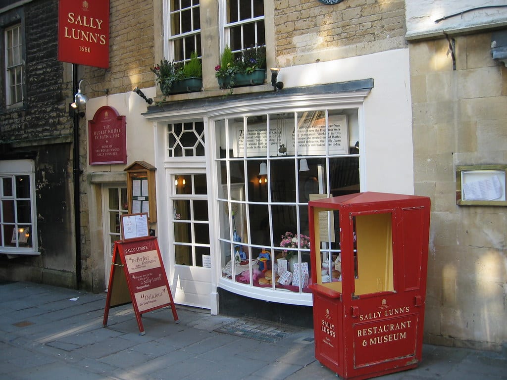 Exterior da Sally Lunn's, uma padaria vermelho e branco com sinalização, com grandes janelas exibindo produtos assados. Vista da rua com fachada histórica de tijolos. Foto para o post de o que fazer em Bath.