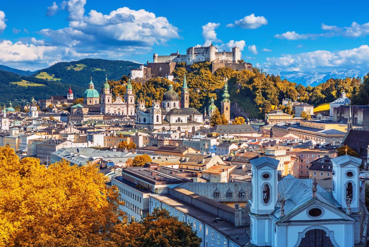 Vista aérea de Salzburgo, Áustria, com um castelo em uma colina, arquitetura barroca e árvores de outono sob um céu azul.