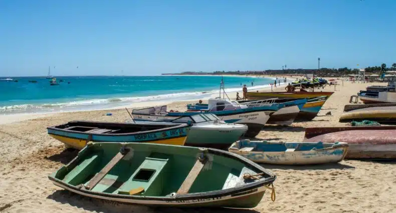 Vários barcos de pesca coloridos em Santa Maria, que pode ser visitado com o eSIM Cabo Verde, repousam em uma praia de areia ao lado de águas azuis e cristalinas sob um céu ensolarado e brilhante.