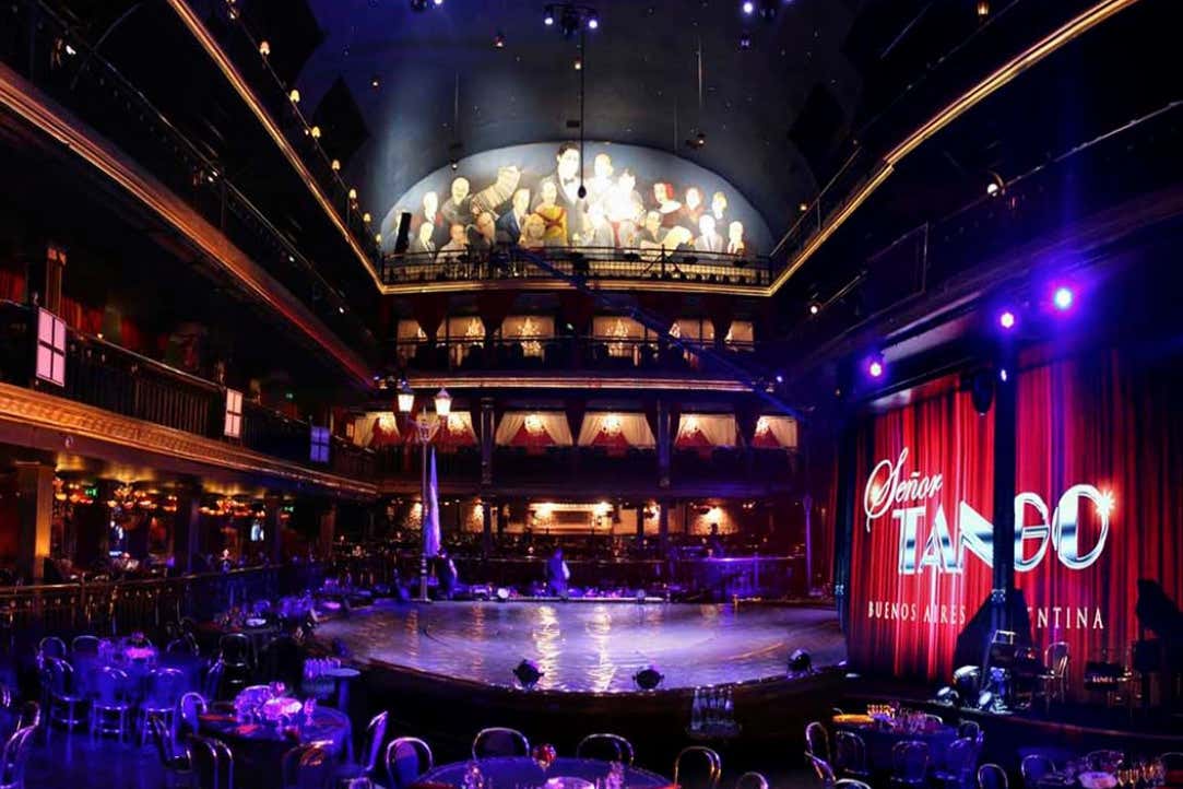 Teatro de tango com pouca iluminação, mesas redondas e um palco, com um mural de pessoas na parede superior e uma cortina vermelha na lateral.
