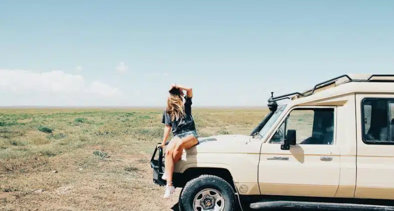 Uma pessoa está sentada no capô de um SUV bege estacionado em uma planície gramada sob um céu azul claro no Serengeti, que pode ser visitado com o eSIM Tanzânia.
