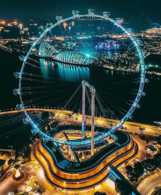 Vista aérea de uma grande roda-gigante iluminada à noite, com uma paisagem urbana e um canal ao fundo.